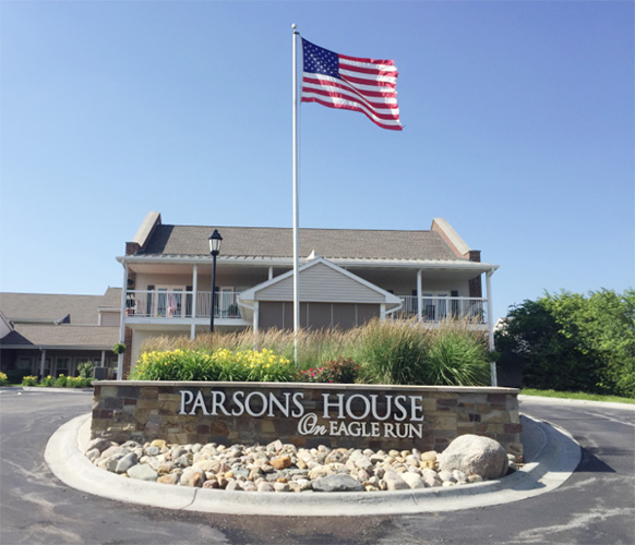 front view of the roundabout in front of Parsons House Assisted Living and Memory Care in Omaha, NE. there is a brick sign with the name and flowers. There is an American flag in the front, too.