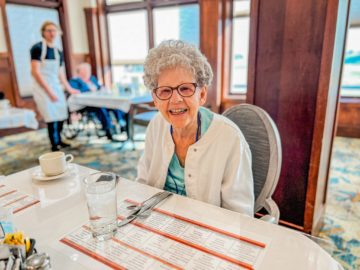 a senior living resident with gray hair, glasses, and a white sweater sitting at a table.